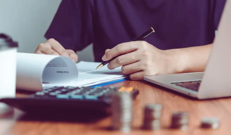 Na zdjęciu widać osobę siedzącą przy drewnianym biurku, zajmującą się dokumentami. Osoba trzyma w jednej ręce długopis, a drugą przewraca strony dokumentu, który leży otwarty na biurku. W tle znajduje się otwarty laptop, sugerujący pracę biurową lub analizę finansową. Na pierwszym planie widoczne są stosy monet oraz kalkulator, co wskazuje na zajęcia związane z finansami, budżetowaniem lub obliczeniami. Atmosfera zdjęcia jest skupiona i profesjonalna, a widok monet i kalkulatora sugeruje, że osoba zajmuje się finansami, rachunkowością lub innymi obliczeniami związanymi z pieniędzmi.