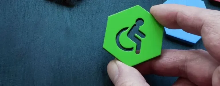 Obrazek wyróżniający do artykułu - symbole niepełnosprawności. Na zdjęciu widzimy dłoń trzymającą sześciokątny, zielony element z wyciętym symbolem osoby na wózku inwalidzkim. Tło jest ciemne, prawdopodobnie granatowe lub czarne, co sprawia, że zielony sześciokąt jest dobrze widoczny. W tle, na górze, można dostrzec inne elementy w różnych kolorach, takie jak czerwony i niebieski, ale są one nieostre i mniej widoczne. Symbol osoby na wózku inwalidzkim jest dobrze wyeksponowany i centralny w kadrze.