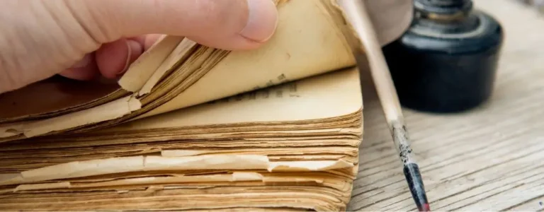 Obrazek wyróżniający do artykułu - mądre cytaty. Na zdjęciu widzimy zbliżenie na starą, pożółkłą książkę o postrzępionych krawędziach stron. Delikatna ręka przewraca jedną ze stron, co wskazuje na ostrożne i pełne szacunku obchodzenie się z tym wiekowym woluminem. Obok książki znajduje się kałamarz z piórem, co sugeruje tradycyjne narzędzia do pisania używane w dawnych czasach. Zdjęcie to emanuje uczuciem nostalgii i szacunku dla literatury oraz rękopiśmiennej sztuki. Przywołuje na myśl czasy, gdy książki były rzadkością i cennymi źródłami wiedzy, a pisanie stanowiło poważne, niemal rytualne zajęcie. Może to wywoływać refleksje nad historią i znaczeniem zachowywania oraz przekazywania wiedzy przez pokolenia.