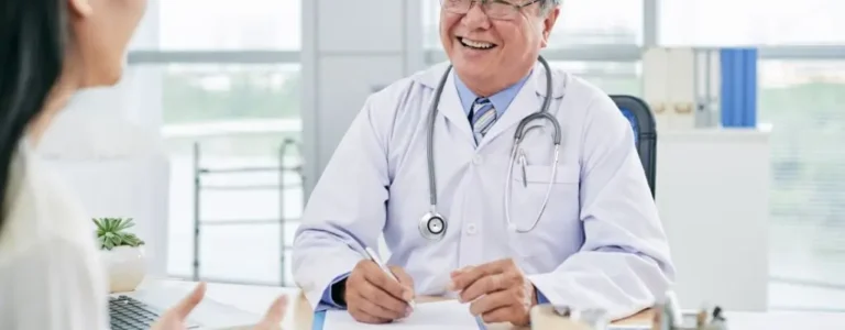 Obrazek wyróżniający do artykułu - zwolnienie lekarskie wstecz. Na zdjęciu widoczny jest starszy lekarz w białym fartuchu, z stetoskopem zawieszonym na szyi, który siedzi przy biurku i uśmiecha się szeroko. Przed nim znajduje się pacjentka, której twarz nie jest w pełni widoczna, ale można zauważyć jej ręce w geście rozmowy. Lekarz wydaje się być w dobrym humorze, co sugeruje pozytywną atmosferę w gabinecie. W tle widać jasne pomieszczenie z dużymi oknami, przez które wpada naturalne światło. Na biurku lekarza znajdują się różne przedmioty, w tym doniczka z rośliną.