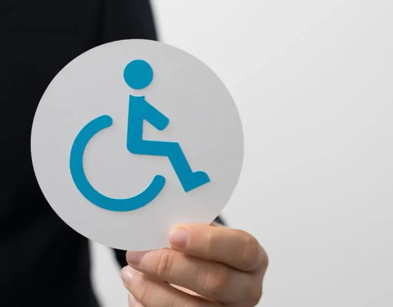 Obrazek wyróżniający do artykułu - ulgi dla niepełnosprawnych. Obraz przedstawia dłoń trzymającą okrągły znak z symbolem osoby niepełnosprawnej na wózku inwalidzkim. Symbol jest niebieski na białym tle. Osoba trzymająca znak ma na sobie ciemne ubranie. Tło jest jasne i jednolite, co sprawia, że symbol jest wyraźnie widoczny. Obraz jest prosty i skupia się na temacie niepełnosprawności oraz związanych z nią ulg i wsparcia.