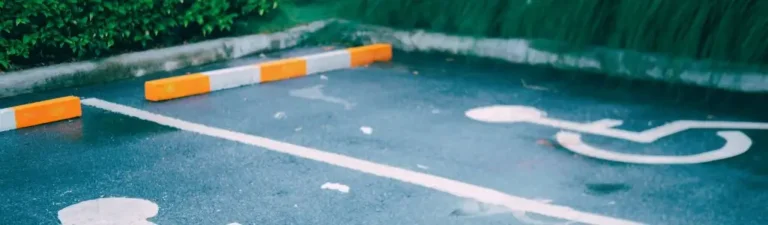 Obrazek wyróżniający do artykułu - miejsce parkingowe dla niepełnosprawnych. Zdjęcie przedstawia miejsce parkingowe dla osób niepełnosprawnych, które jest oznaczone dużą, białą ikoną wózka inwalidzkiego namalowaną na ciemnoszarej, asfaltowej nawierzchni. Miejsce to znajduje się na zewnątrz, prawdopodobnie w pobliżu jakiegoś budynku lub obiektu publicznego. W tle widać zielony żywopłot, który dodaje otoczeniu nieco zieleni. Na granicy miejsca parkingowego widoczne są biało-pomarańczowe odbojnice. Cała powierzchnia parkingu jest mokra, co sugeruje, że zdjęcie zostało zrobione po deszczu lub w wilgotny dzień.