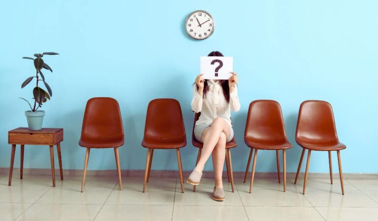 Obrazek wyróżniający do artykułu - słabe strony podczas rozmowy kwalifikacyjnej. Na obrazie znajduje się kobieta siedząca na jednym z pięciu brązowych krzeseł ustawionych w rzędzie pod ścianą w jasnoniebieskim kolorze. Kobieta jest ubrana w kremową sukienkę i beżowe buty na koturnie. Trzyma przed sobą kartkę papieru z dużym, czarnym znakiem zapytania, zasłaniając nią swoją twarz. Nad krzesłami, na ścianie, wisi okrągły, klasyczny zegar wskazujący godzinę 11:05. Po lewej stronie kadru, obok krzeseł, znajduje się drewniany stolik z rośliną doniczkową o dużych, ciemnozielonych liściach.