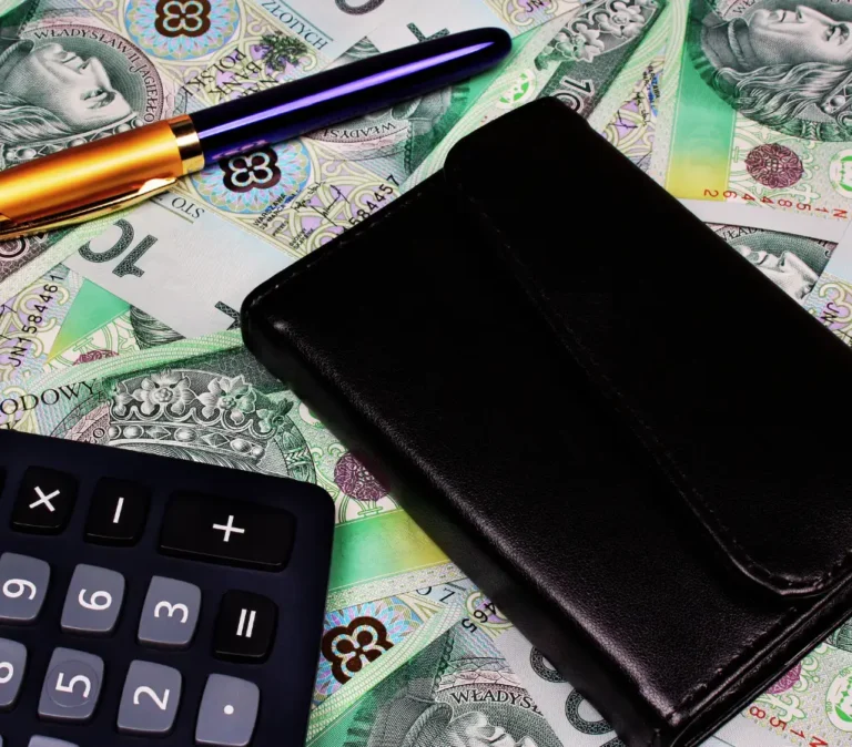 Obrazek wyróżniający do artykułu - ile pracodawca dostaje za pracownika z grupą. Obrazek przedstawia zestaw przedmiotów na tle polskich banknotów (w tym o nominałach 100 zł), sugerując tematykę związaną z finansami. Na banknotach znajdują się elementy w kolorach zielonym, fioletowym i złotym. Na pierwszym planie widać czarny portfel, długopis o złoto-granatowej obudowie oraz kalkulator z widocznymi cyframi i podstawowymi funkcjami matematycznymi. Cała scena wskazuje na kontekst związany z zarządzaniem finansami, takimi jak liczenie pieniędzy lub planowanie wydatków.