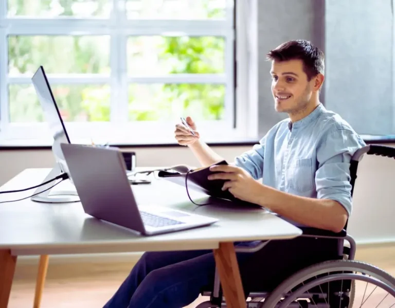 Obrazek wyróżniający do artykułu - uprawnienia pracowników z orzeczeniem. Na zdjęciu widoczny jest młody mężczyzna siedzący na wózku inwalidzkim przy biurku w jasnym, nowoczesnym biurze. Mężczyzna uśmiecha się, patrząc w stronę swojego komputera, a w dłoniach trzyma notes, co sugeruje, że jest zaangażowany w pracę lub spotkanie online. Na biurku znajdują się dwa laptopy, co wskazuje na intensywną pracę lub wykonywanie kilku zadań jednocześnie. W tle widać duże okno, przez które wpada naturalne światło, oświetlając przestrzeń roboczą. Zdjęcie pokazuje osobę z niepełnosprawnością w profesjonalnym środowisku, co podkreśla integrację oraz aktywne uczestnictwo w pracy biurowej.