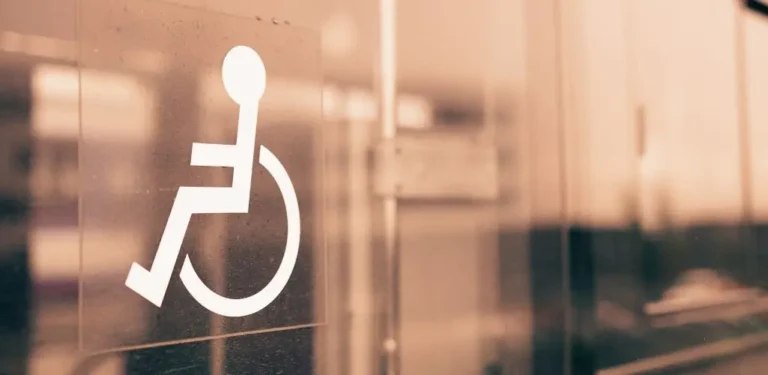 Obrazek wyróżniający do artykułu - co daje lekki stopień niepełnosprawności. Na zdjęciu znajduje się symbol osoby na wózku inwalidzkim, umieszczony na szklanej powierzchni, prawdopodobnie na drzwiach lub oknie. Symbol jest biały i wyraźnie widoczny na tle szkła, które odbija delikatne, ciepłe światło w odcieniach sepii. Zdjęcie przedstawia dostępność przestrzeni publicznej dla osób z niepełnosprawnościami, wskazując na miejsce dostosowane do ich potrzeb. Może być związane z tematyką udogodnień, praw lub świadomości społecznej dotyczącej osób z niepełnosprawnościami.