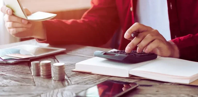 Obrazek wyróżniający do artykułu - świadczenia dla niepełnosprawnych. Obraz przedstawia osobę w czerwonym swetrze siedzącą przy drewnianym stole. Na stole znajdują się monety ustawione w stosiki, kalkulator, notatnik oraz urządzenia elektroniczne, takie jak tablet i telefon. Osoba przelicza coś na kalkulatorze, trzymając długopis w dłoni. Scena nawiązuje do zarządzania finansami lub obliczania świadczeń.