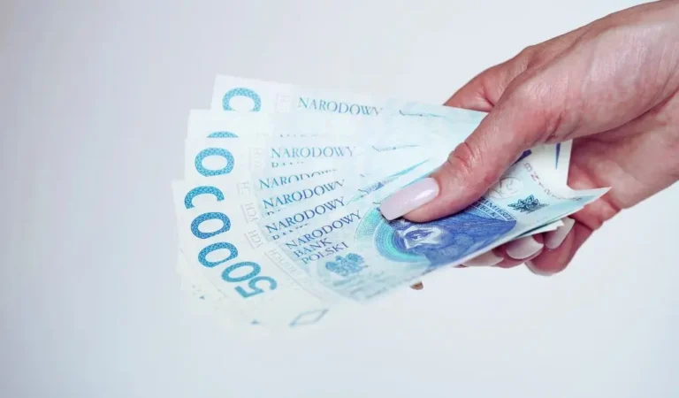 Na zdjęciu widoczna jest kobieca dłoń z zadbanymi, jasnymi paznokciami, która trzyma wachlarz banknotów o nominale 500 zł. Banknoty są wyraźnie oznaczone napisem „Narodowy Bank Polski” i przedstawiają postać historyczną na awersie. Całość wywołuje odczucie stabilności finansowej, pewności siebie i komfortu. Obrazek symbolizuje potencjalny dodatek dopełniający do budżetu lub wydatków.