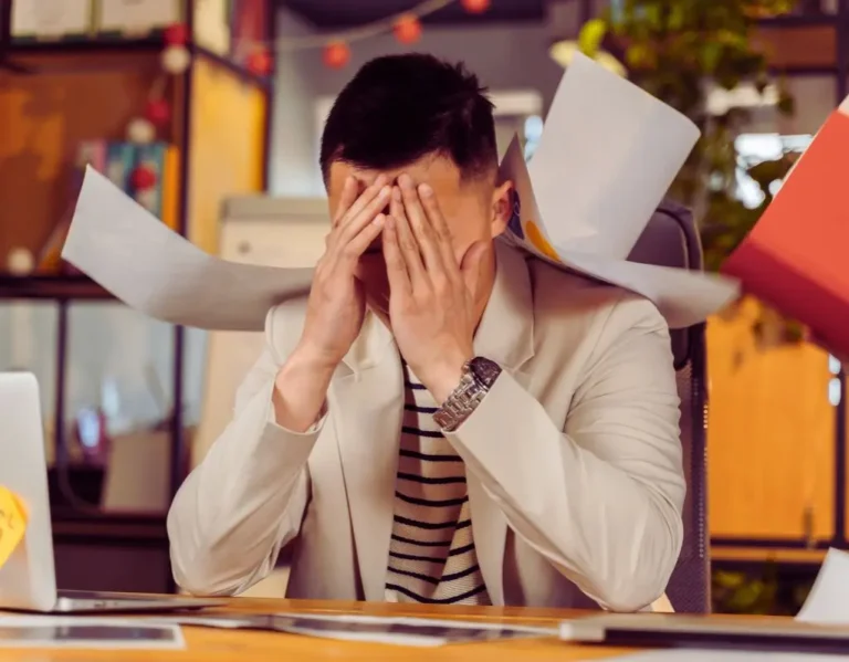 Na obrazie widzimy mężczyznę siedzącego przy biurku w biurze, wyraźnie przytłoczonego i zestresowanego. Jest ubrany w jasną marynarkę i koszulkę w paski. Zakrywa twarz dłońmi, co sugeruje frustrację, zmęczenie lub bezsilność. Wokół niego znajdują się porozrzucane dokumenty, które spadają na jego głowę i ramiona, symbolizując chaos i presję w pracy. W tle widoczne są elementy biurowe, takie jak rośliny i regały, które kontrastują z emocjonalnym napięciem przedstawionej sytuacji. Obraz w sposób wymowny ilustruje problem, jak radzić sobie z mobbingiem w pracy, wywołując skojarzenia z przeciążeniem, stresem i poczuciem osamotnienia w trudnym środowisku zawodowym.