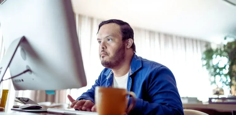 Na zdjęciu znajduje się mężczyzna z zespołem Downa, który siedzi przed komputerem, skupiony na pracy. Ma na sobie niebieską koszulę i patrzy na ekran z wyrazem pełnego zaangażowania i koncentracji. W tle widać jasne zasłony oraz rozmytą zieloną roślinę, co nadaje wnętrzu przytulny charakter. Na pierwszym planie stoi pomarańczowy kubek, który dodaje ciepłego akcentu do sceny. Obraz ukazuje osobę z niepełnosprawnością, która w pełni wykorzystuje swoje umiejętności, pokazując, że różne stopnie niepełnosprawności nie ograniczają zdolności do efektywnego działania i realizowania się w pracy. Scena emanuje poczuciem determinacji i wrażeniem, że każdy człowiek może osiągnąć swoje cele, niezależnie od okoliczności.