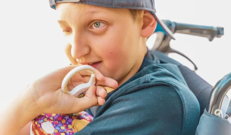 Na zdjęciu widać chłopca siedzącego na wózku inwalidzkim. Ma na sobie ciemną koszulkę i czapkę z daszkiem założoną tyłem. Jego jasne oczy błyszczą w świetle, a twarz wyraża ciekawość i radość. Trzyma w dłoni zabawkę w kształcie kółka, którą ktoś mu podaje – to gest pełen troski i bliskości. Zdjęcie oddaje ciepłą atmosferę i autentyczne emocje. Pokazuje codzienność osób z Niepełnosprawność sprzężona to nie tylko wyzwania, ale także radość, wsparcie i relacje z bliskimi. Jasne światło i miękkie kolory dodają tej scenie spokoju i pozytywnej energii.