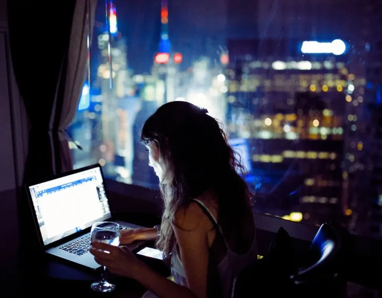 Kobieta pracuje w nocy przy laptopie, siedząc przy oknie z widokiem na rozświetlone miasto. W jednej dłoni trzyma kieliszek z wodą, a drugą obsługuje telefon. Blask ekranu oświetla jej twarz, kontrastując z ciemnością wnętrza. Za szybą rozpościera się panorama nowoczesnych wieżowców z migającymi światłami. Zdjęcie oddaje atmosferę skupienia i samotności – nocną ciszę przerywa jedynie rytm pracy i pulsujące światła miasta. Praca w nocy osoby niepełnosprawnej może być zarówno wyzwaniem, jak i momentem spokoju, gdy świat zwalnia, a obowiązki wypełniają przestrzeń ciszy.