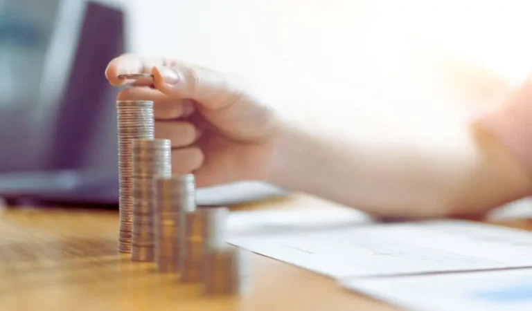 Na zdjęciu widzimy dłoń układającą monety w rosnące stosy na drewnianym biurku. Monety są starannie ułożone, a największy stos jest właśnie powiększany o kolejną monetę. W tle znajduje się rozmyty laptop i dokumenty, co sugeruje kontekst związany z finansami, oszczędzaniem lub budżetowaniem. Zdjęcie oddaje poczucie dbałości o finanse, planowania przyszłości i świadomego zarządzania pieniędzmi. Może kojarzyć się z trudami oszczędzania, ale też z satysfakcją płynącą z kontrolowania własnych dochodów i wydatków. W kontekście tematu „ile zarabia osoba z orzeczeniem” obrazek symbolizuje stopniowe budowanie stabilności finansowej i znaczenie każdej złotówki w budżecie osoby z orzeczeniem o niepełnosprawności.