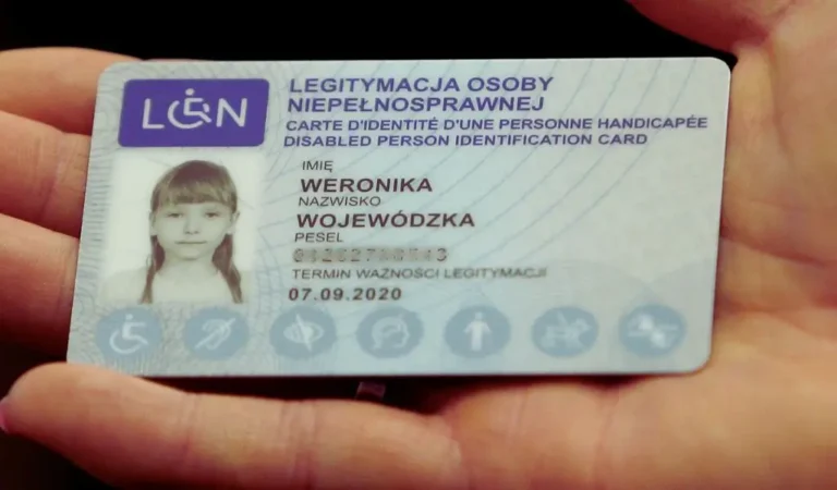 Na zdjęciu widoczna jest Legitymacja osoby niepełnosprawnej, trzymana w dłoni. Dokument ma jasnoniebieskie tło z subtelnymi wzorami i symbolami wskazującymi na różne rodzaje niepełnosprawności. W lewym górnym rogu znajduje się niebieski znak z symbolem wózka inwalidzkiego i literami "LON". W centralnej części znajduje się czarno-białe zdjęcie młodej dziewczynki o jasnych włosach, z grzywką, patrzącej spokojnym wzrokiem. Dane osobowe, takie jak imię, nazwisko oraz PESEL (częściowo ukryty), są wydrukowane w wyrazistej czcionce. Poniżej znajduje się termin ważności dokumentu, który upłynął 7 września 2020 roku.