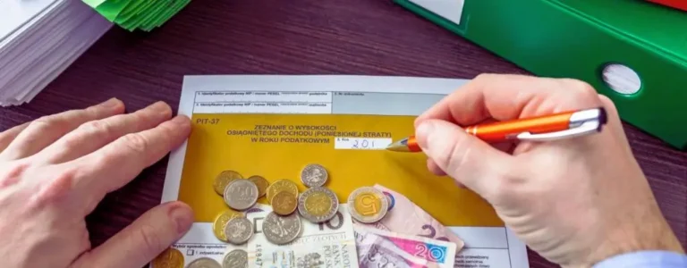 Na zdjęciu widać ręce osoby wypełniającej formularz PIT-37, który służy do rozliczenia podatkowego. Dokument jest częściowo przykryty polskimi banknotami i monetami, symbolizującymi kwestie finansowe, takie jak zwrot za leki dla niepełnosprawnych czy inne ulgi podatkowe. Osoba trzyma długopis i wpisuje rok podatkowy, co sugeruje moment rozliczenia – często pełen napięcia i refleksji nad finansami. W tle znajdują się kolorowe segregatory i stosy dokumentów, podkreślające urzędowy charakter sytuacji. Zdjęcie oddaje uczucie skupienia i odpowiedzialności, które towarzyszą wypełnianiu formalności podatkowych, szczególnie w kontekście ważnych ulg i zwrotów.