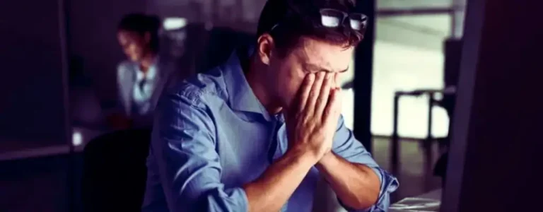 Mężczyzna w biurze, ubrany w niebieską koszulę, siedzi przed komputerem i trzyma twarz w dłoniach w geście zmęczenia lub frustracji. Na głowie ma okulary, a w tle widać inną osobę przy biurku, co sugeruje pracę w późnych godzinach. Obraz oddaje poczucie wyczerpania, przytłoczenia i stresu związanego z pracą po zmroku. Idealny do zilustrowania artykułu o temacie jak radzić sobie z nocną zmianą – ukazuje zarówno fizyczne, jak i emocjonalne skutki pracy nocą.
