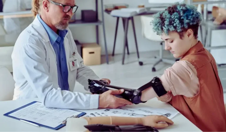 Na zdjęciu widzimy młodą osobę z niepełnosprawnością, która podczas konsultacji z protetykiem przymierza nowoczesną, czarną protezę ręki. Specjalista w białym fartuchu z troską i zaangażowaniem pomaga jej w dopasowaniu urządzenia. Na stole leżą dokumenty oraz inne modele protez. W tle widoczne jest wnętrze gabinetu protetycznego – przestrzeń profesjonalna, ale też spokojna i wspierająca. Obraz oddaje moment skupienia, nadziei i wsparcia, pokazując, jak ważną rolę odgrywa pomoc specjalistów i uzyskane z PFRON dofinansowanie do protezy kończyny w procesie powrotu do samodzielności.