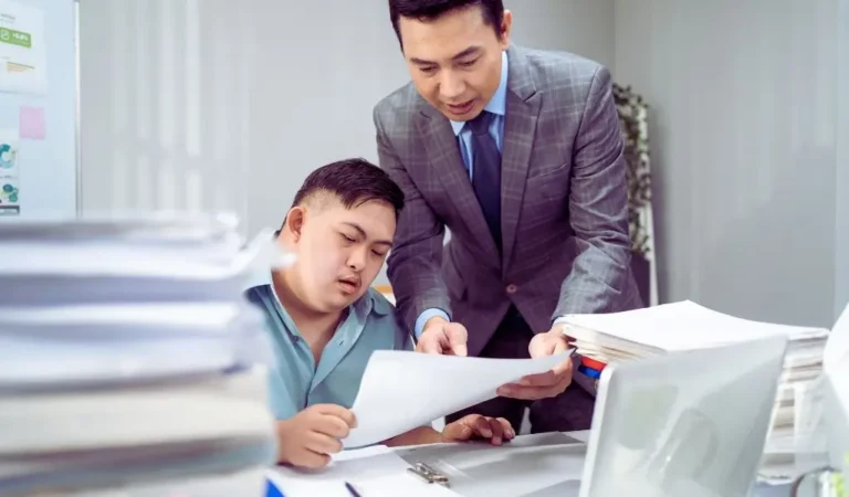 Na zdjęciu widzimy dwóch mężczyzn pracujących razem przy biurku w biurze pełnym dokumentów. Jeden z nich to młody pracownik z zespołem Downa, który siedzi i skupia się na przeglądaniu dokumentu. Obok niego stoi jego przełożony w garniturze, który pochyla się i z zaangażowaniem coś wyjaśnia, wskazując na kartkę. Panuje atmosfera zaufania, wsparcia i współpracy. Obrazek oddaje moment, który może symbolizować sytuację, gdy pracownik przynosi orzeczenie o niepełnosprawności – pokazuje otwartość, profesjonalizm i szacunek w miejscu pracy.