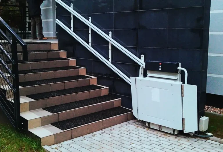 Na zdjęciu widać zewnętrzne schody prowadzące do budynku, przy których zamontowano platformę przyschodową — specjalistyczne urządzenie umożliwiające osobom poruszającym się na wózkach inwalidzkich pokonanie barier poziomych. Platforma jest złożona i gotowa do użycia, a jej prowadnice biegną wzdłuż poręczy zamocowanej do ściany. Całość prezentuje się solidnie i nowocześnie, a starannie ułożony chodnik pod platformą podkreśla dbałość o detale. Obrazek oddaje konkretne, pozytywne emocje — poczucie dostępności, szacunku i troski o drugiego człowieka. Pokazuje, że likwidacja barier architektonicznych z dofinansowaniem PFRON to nie tylko techniczne rozwiązanie, ale realna zmiana w życiu osób z niepełnosprawnością – na lepsze, bardziej niezależne i godne.