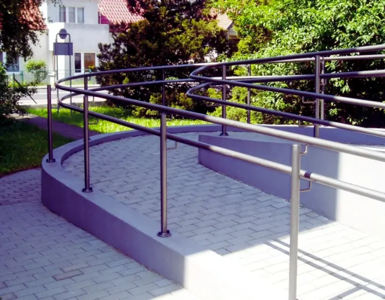Na zdjęciu znajduje się elegancko zaprojektowany podjazd dla niepełnosprawnych, wykonany z jasnej kostki brukowej i otoczony solidną balustradą ze stali nierdzewnej. Konstrukcja łagodnie wije się w górę, tworząc płynne, zakrzywione przejście. Otoczenie jest zielone i spokojne – w tle widać drzewa, krzewy i budynki mieszkalne. Całość sprawia wrażenie dostępnej i przyjaznej przestrzeni. Podjazd dla niepełnosprawnych symbolizuje tutaj troskę o równość, swobodę poruszania się i dbałość o komfort osób z ograniczoną mobilnością.